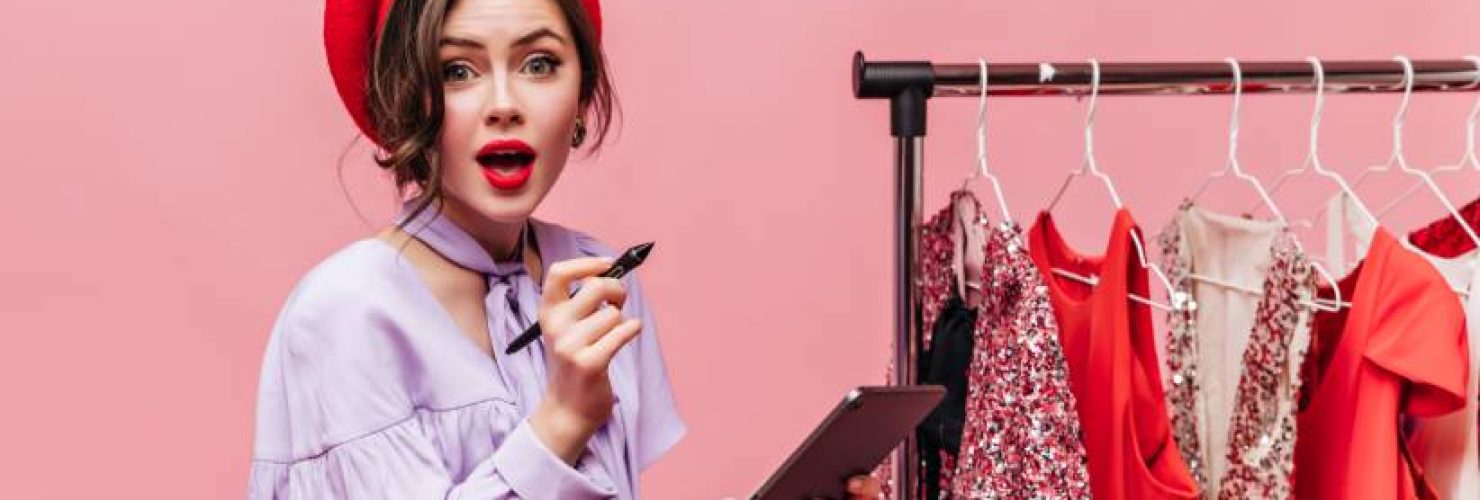 green-eyed-lady-red-hat-with-red-lips-is-holding-tablet-looking-camera-against-background-hangers-with-clothes-1